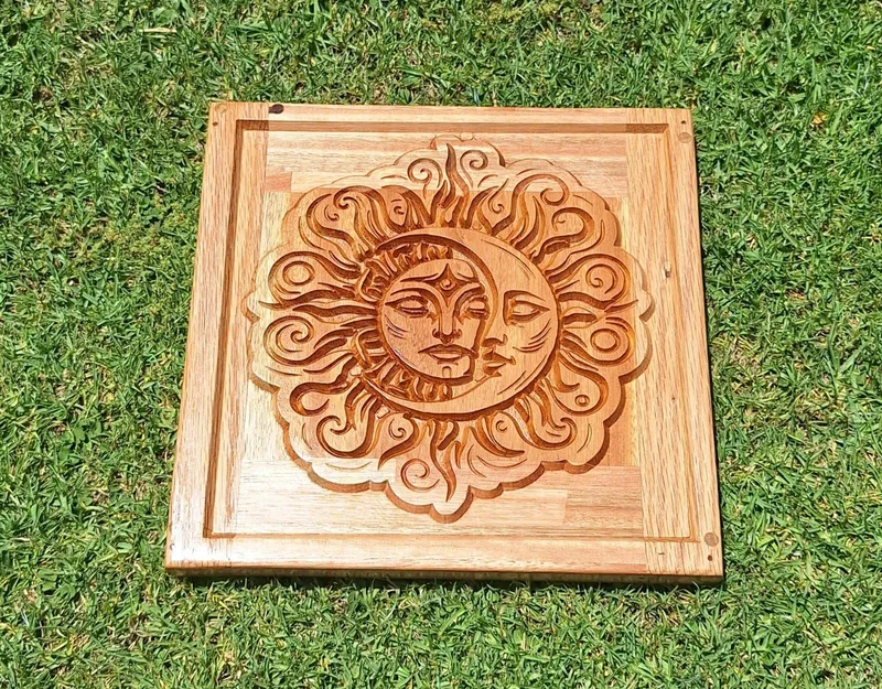Square wooden wall panel carved in relief with a joined sun and crescent moon face inside a plain frame.
