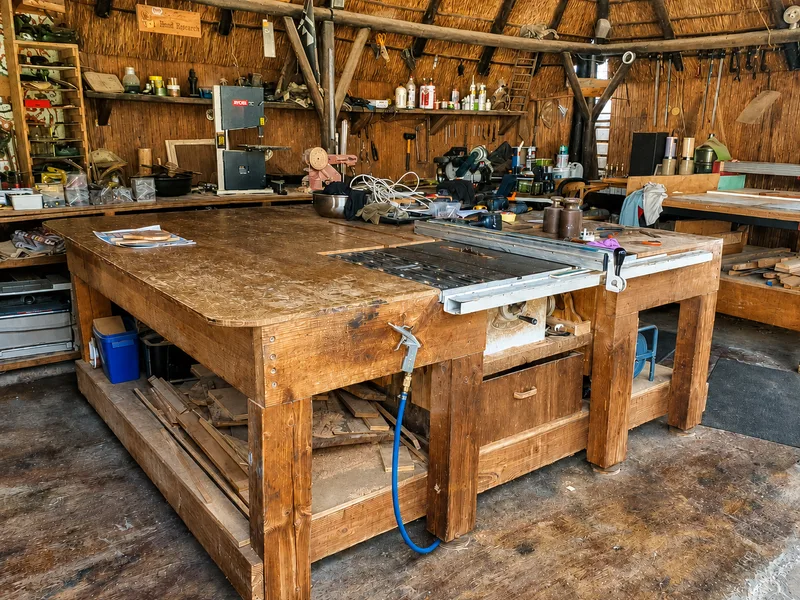 Large timber workbench with a built-in table saw inside a thatched workshop, tools on the walls behind.