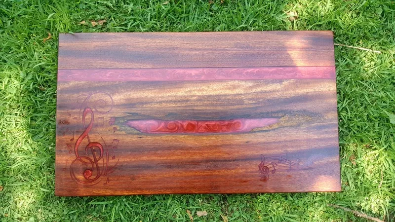 Rectangular dark wooden board with pink resin-filled cracks and burned-in treble clef and music notes.