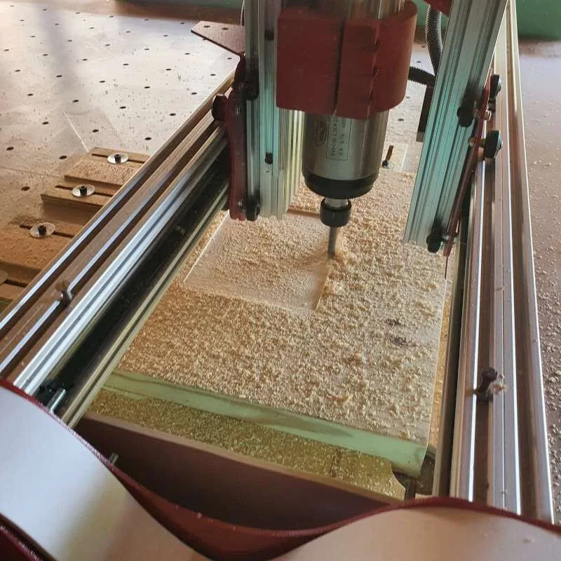 A CNC router spindle cutting into a clamped wooden board covered in sawdust.