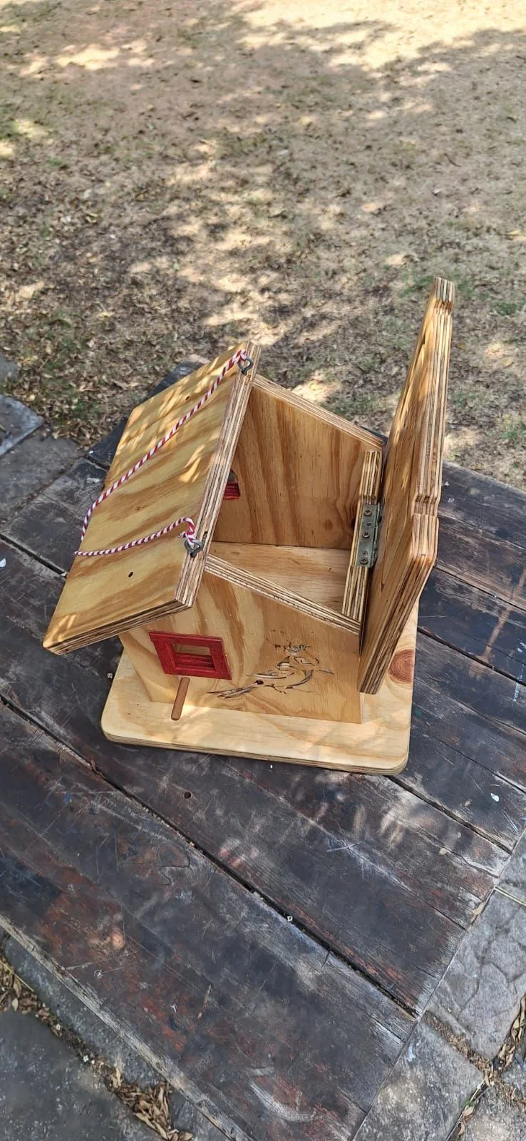 Plywood birdhouse with its hinged roof panel open, showing a red window, dowel perch and burnt bird motif.