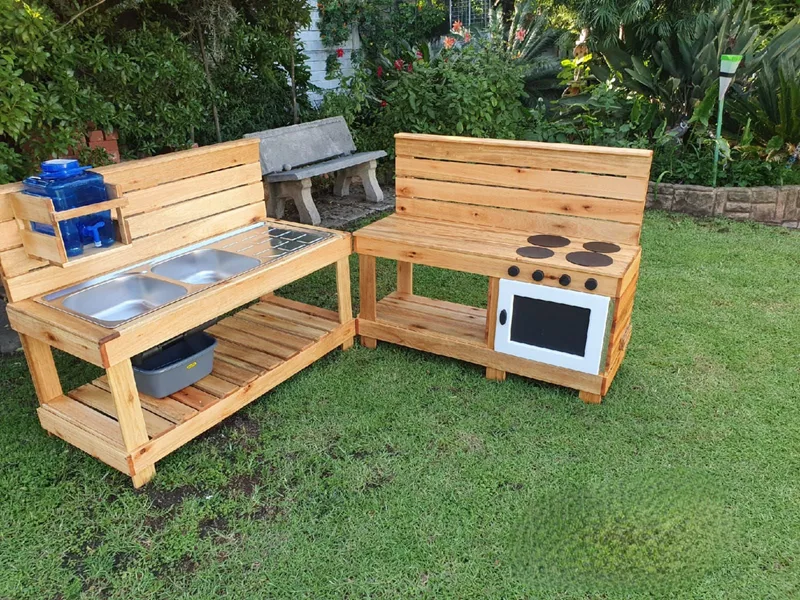 L-shaped wooden mud kitchen with double stainless sink, water bottle holder, painted oven and hob.