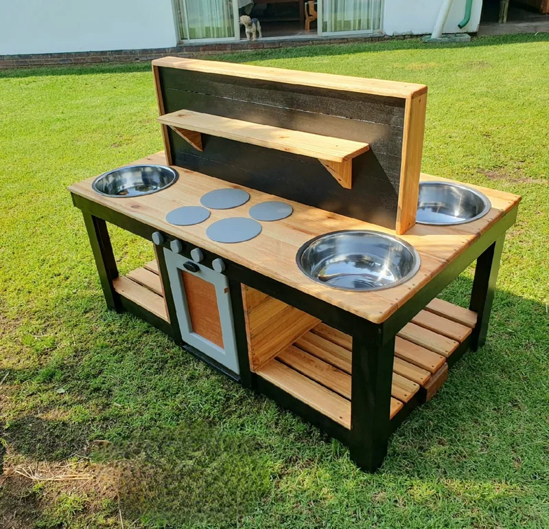 Wooden mud kitchen with three steel bowls, painted hobs, oven door, chalkboard back and lower slatted shelves.
