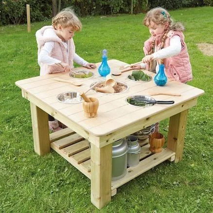 Square wooden outdoor play table with a slatted lower shelf, used by two children with bowls and pans.
