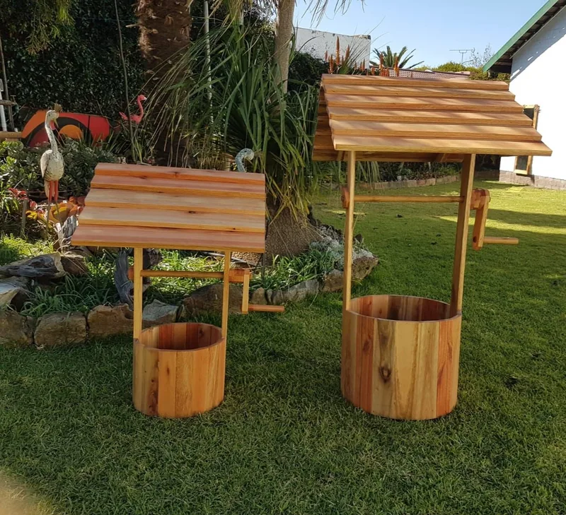 Two timber wishing wells of different sizes on a lawn, each with a round barrel and pitched slatted roof.