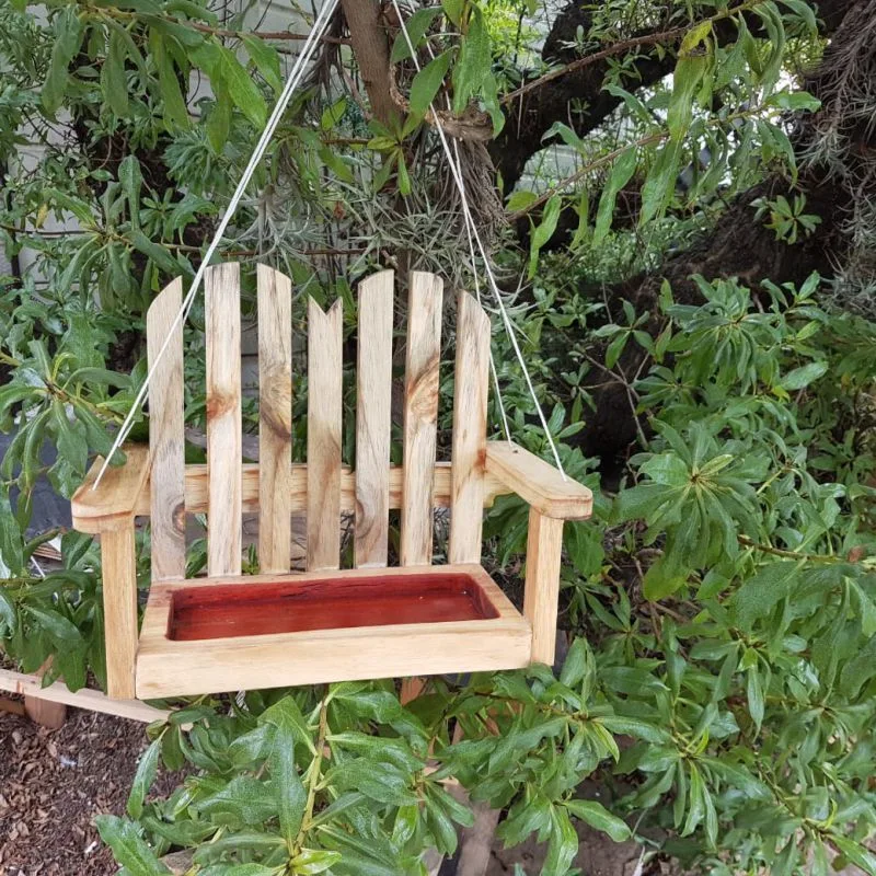 Small wooden bench-shaped bird feeder with slatted back and red-finished seed tray, hung by cord from a tree.
