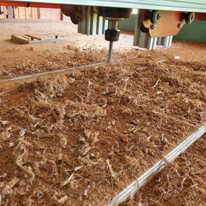 CNC router bit cutting a wooden board on a workshop bed covered in sawdust and shavings.