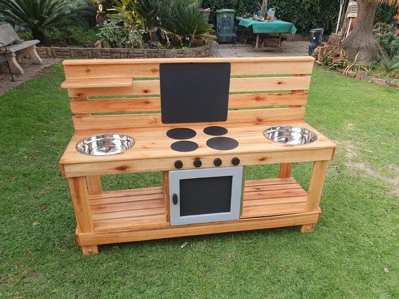 Wooden mud kitchen with two stainless steel bowls, painted hob, oven door, chalkboard and lower shelf.