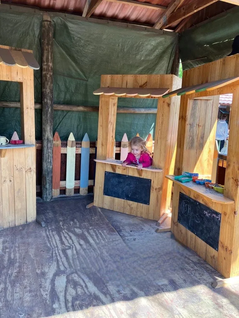 Wooden play shop stall with serving counter, scalloped awning and black chalkboard front panel, child leaning over.