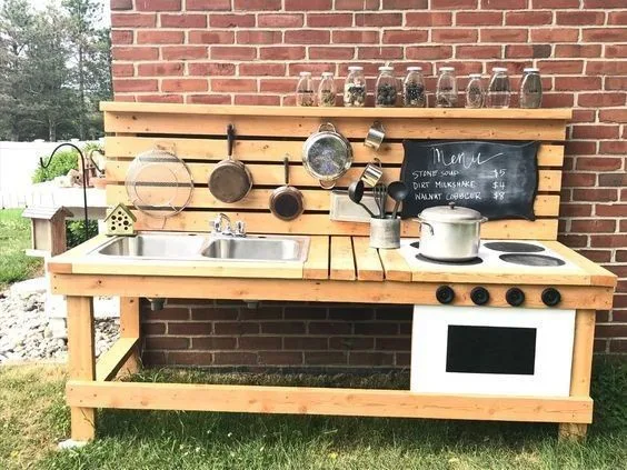 Long timber mud kitchen with two metal sinks, painted oven with knobs, hanging pots and a chalkboard menu.