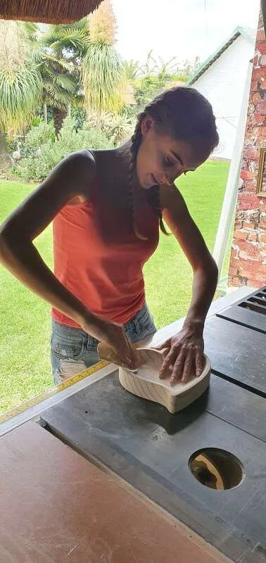 A young woman shaping a small curved wooden block by hand on a saw table in an outdoor workshop.