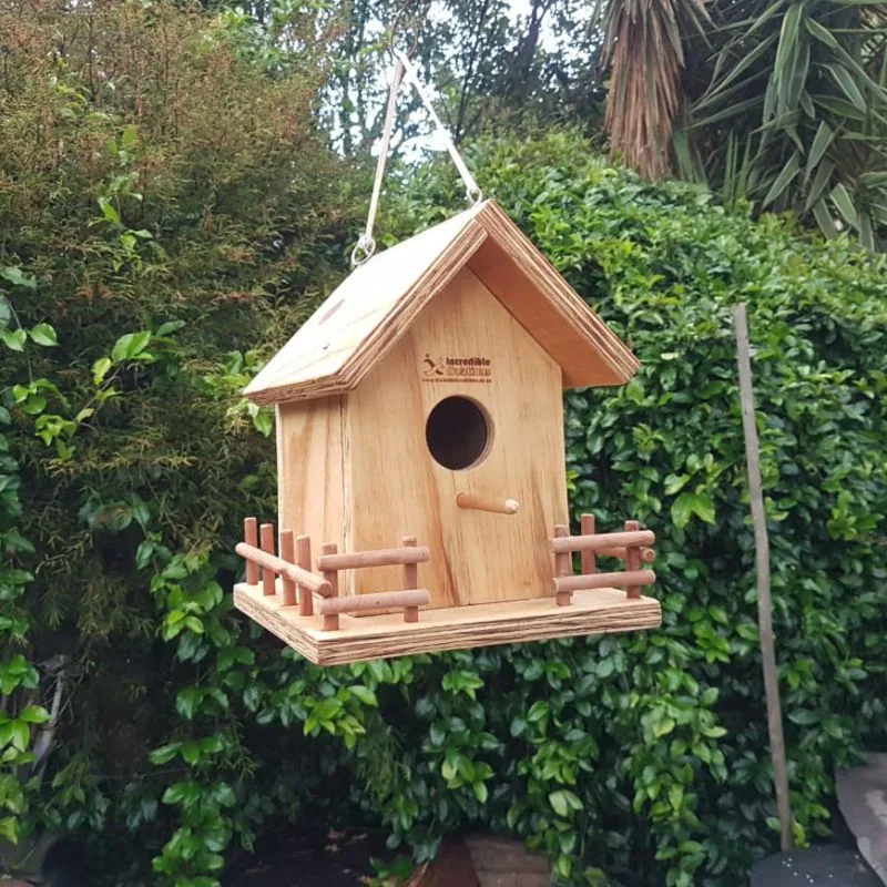 Wooden bird house with pitched roof, round hole, dowel perch and small fenced veranda, hanging from a rope.