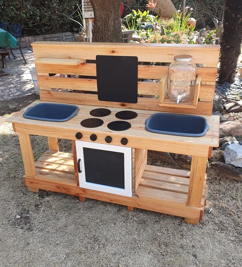 Wooden double mud kitchen with two blue basins, painted hob, oven door, slatted shelves and a glass water dispenser.