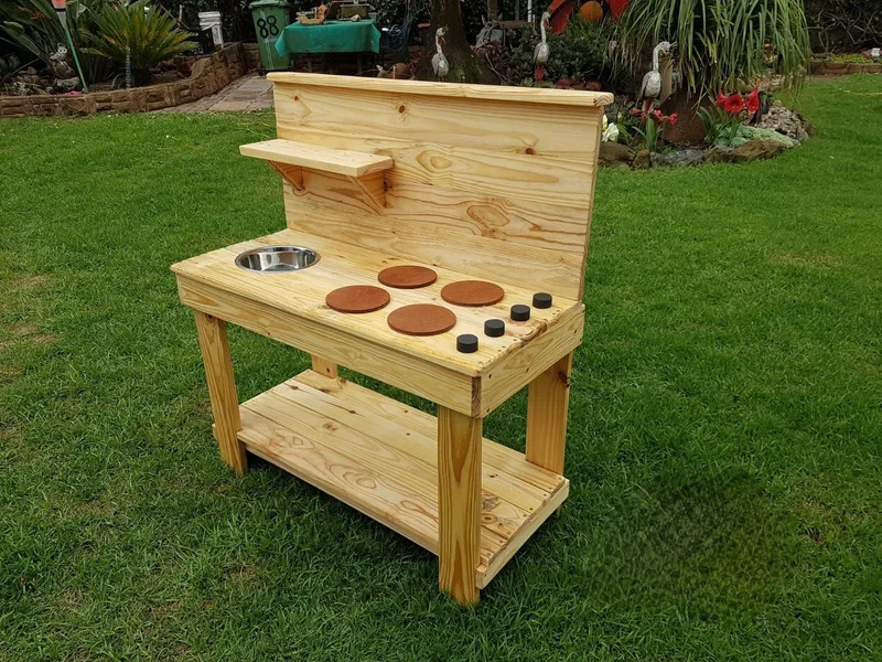 Wooden mud kitchen with a round steel sink bowl, four painted hob plates, upper shelf and lower slatted shelf.