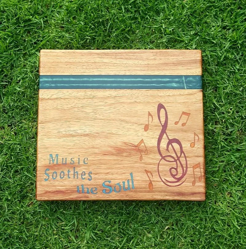 Square wooden cutting board with a teal resin stripe, a treble clef, music notes and the words "Music Soothes the Soul".