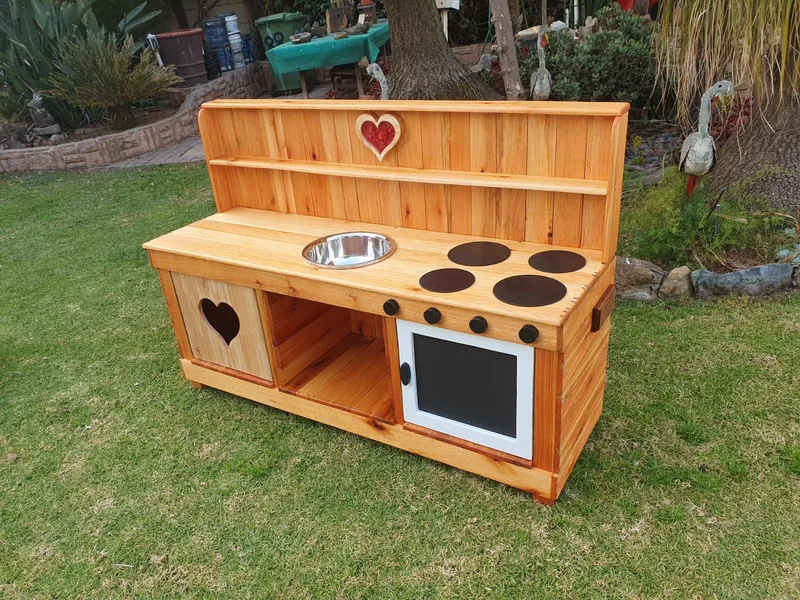 Wooden children's mud kitchen with steel sink bowl, four painted hob plates, oven door and heart cut-outs.